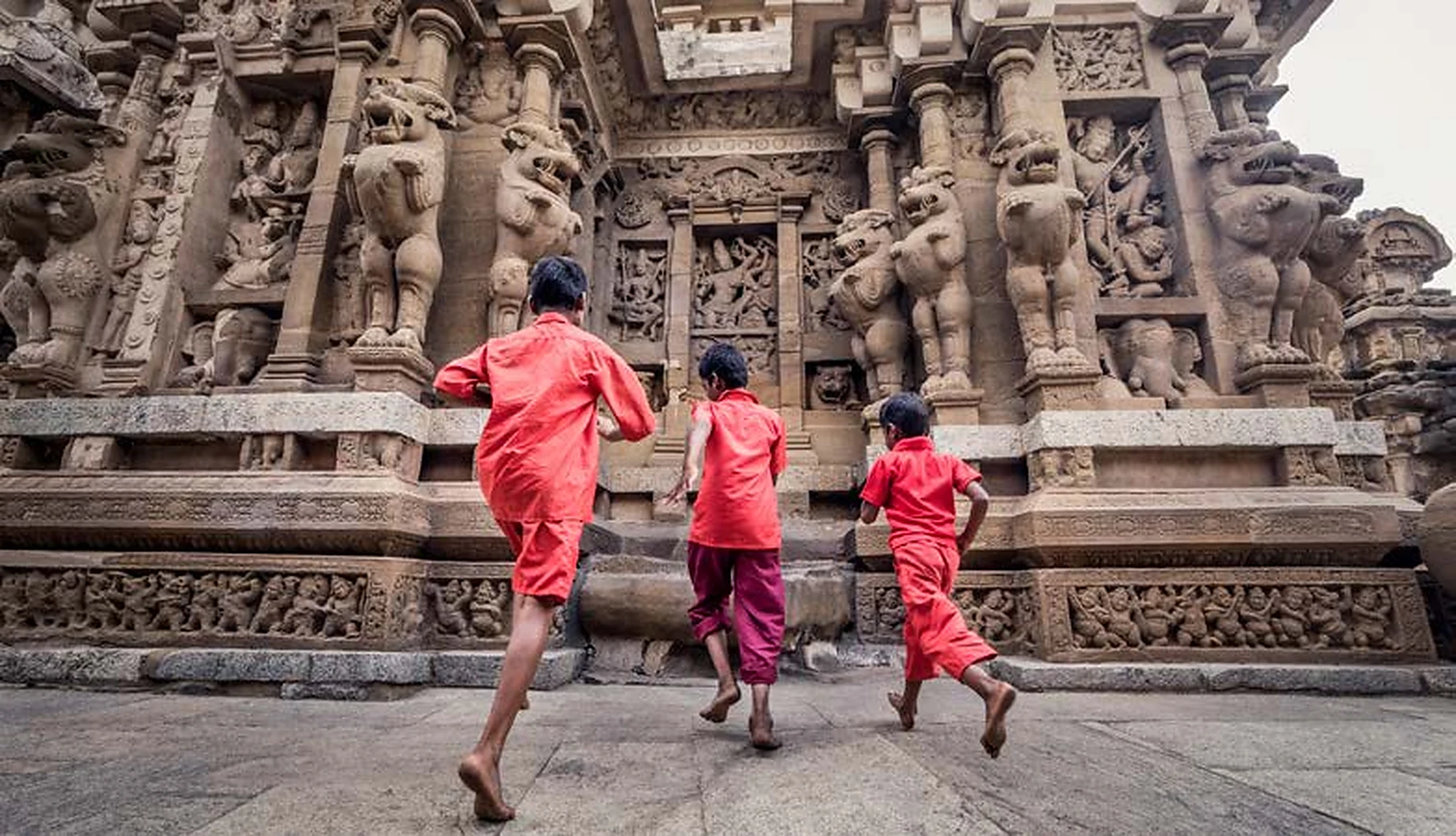 Three children in red clothing running toward a richly carved temple