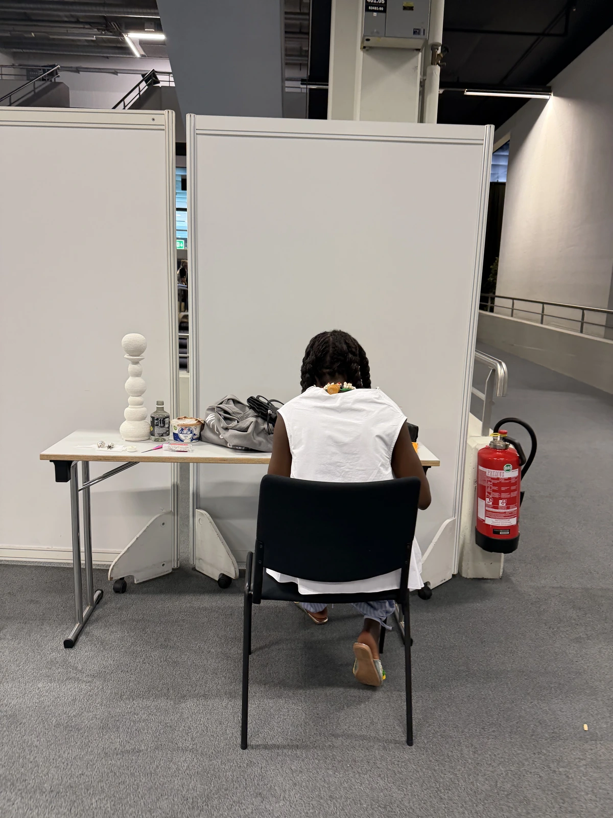 Luma Nascimento seen from behind, working alone at a folding table behind the booth partitions, a white stacked sphere sculpture beside her