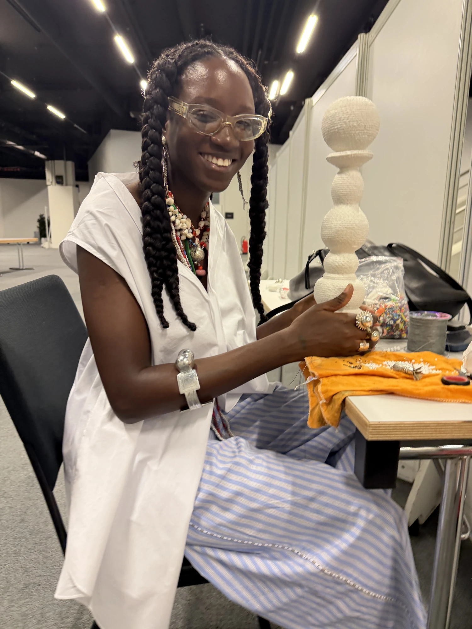 Luma Nascimento smiling at her work table, holding upright a tall white sculpture of stacked wrapped spheres