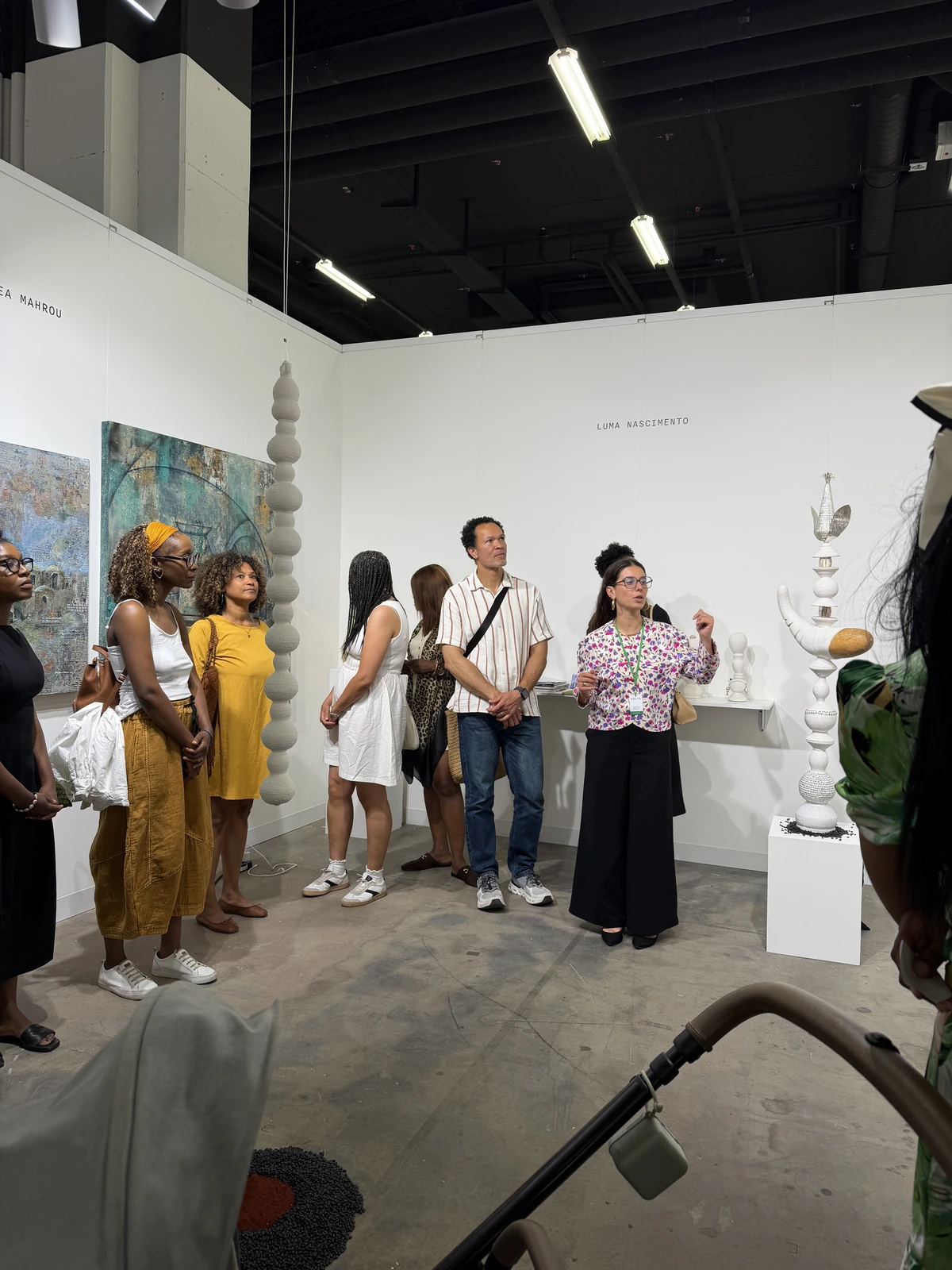 The booth at VOLTA with Luma Nascimento's name on the wall, hanging columns of stacked grey spheres and sculptures on plinths, visitors listening to a walkthrough