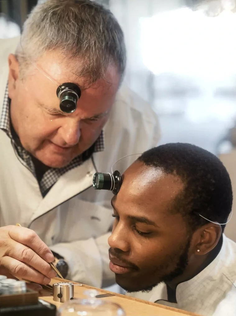 A senior watchmaker and a younger colleague, both wearing loupes, working together over a movement at a watchmaker's bench